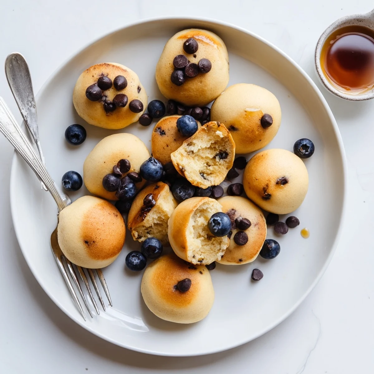 Golden-brown Pancake Poppers with fluffy centers arranged on a wooden board beside maple syrup drizzle and fresh blueberries.
