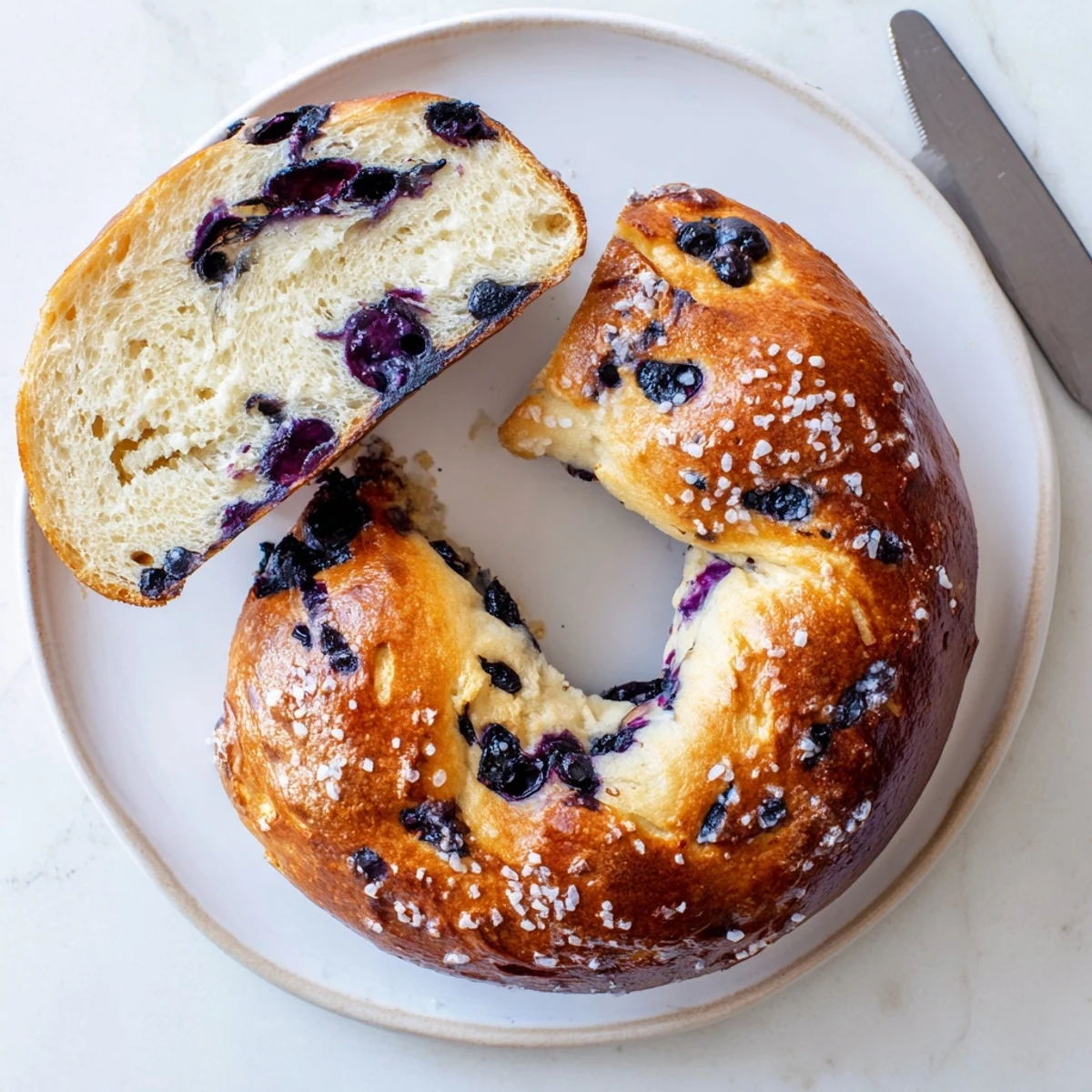 Two toasted gluten-free blueberry bagels with cream cheese spread, served on a plate with fresh blueberries nearby.