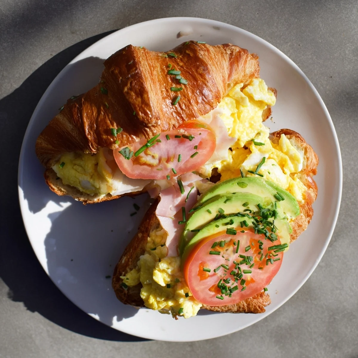 Serving flaky croissant breakfast sandwich with tomato and chives beside a steaming coffee mug.