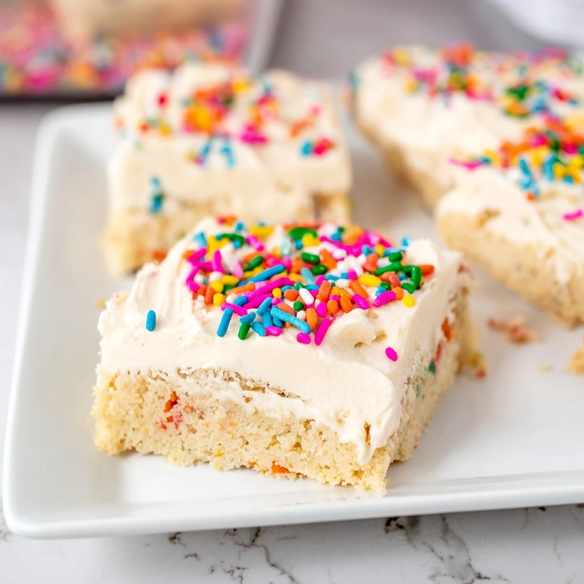 Soft golden sugar cookie bars topped with fluffy white frosting and colorful sprinkles on a white plate
