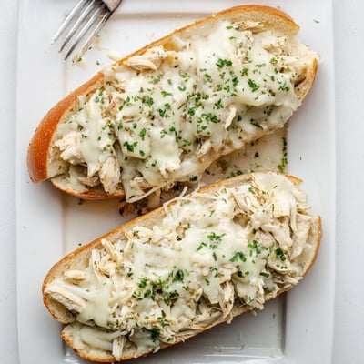 A close-up of warm Viral Chicken Alfredo Garlic Bread reveals bubbly cheese, garlic butter, and shredded chicken on a crusty baguette half.